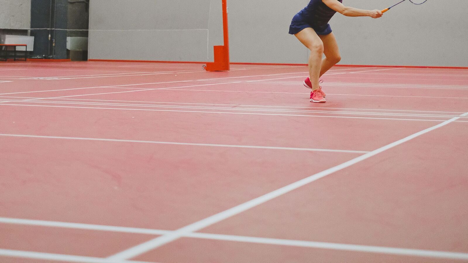 Une personne qui joue au badminton fait une fente vers le filet pour jouer un coup sur un terrain intérieur marqué, avec le poteau du filet et les lignes de service et de côté qui se croisent visibles sur la surface rouge