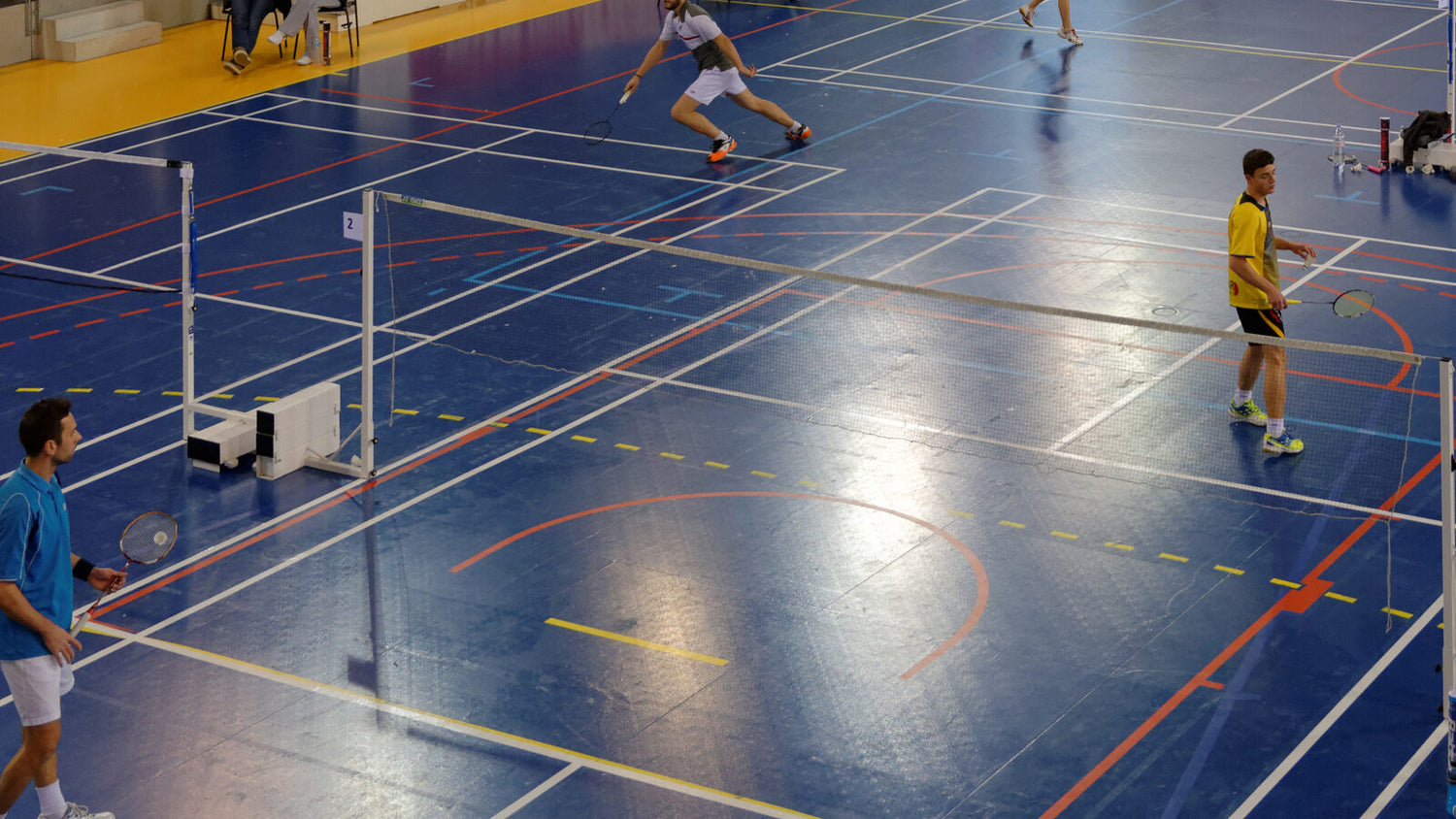 Des personnes jouent au badminton sur plusieurs terrains dans un gymnase intérieur; deux sont au filet et une autre se fend pour atteindre un coup.