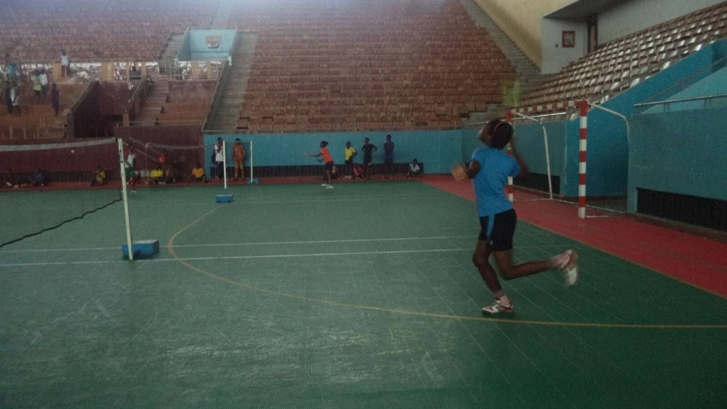 Une jeune joueuse s’élance pour effectuer un smash au-dessus de la tête sur un terrain de badminton intérieur, avec les poteaux du filet et des gradins vides visibles derrière elle.