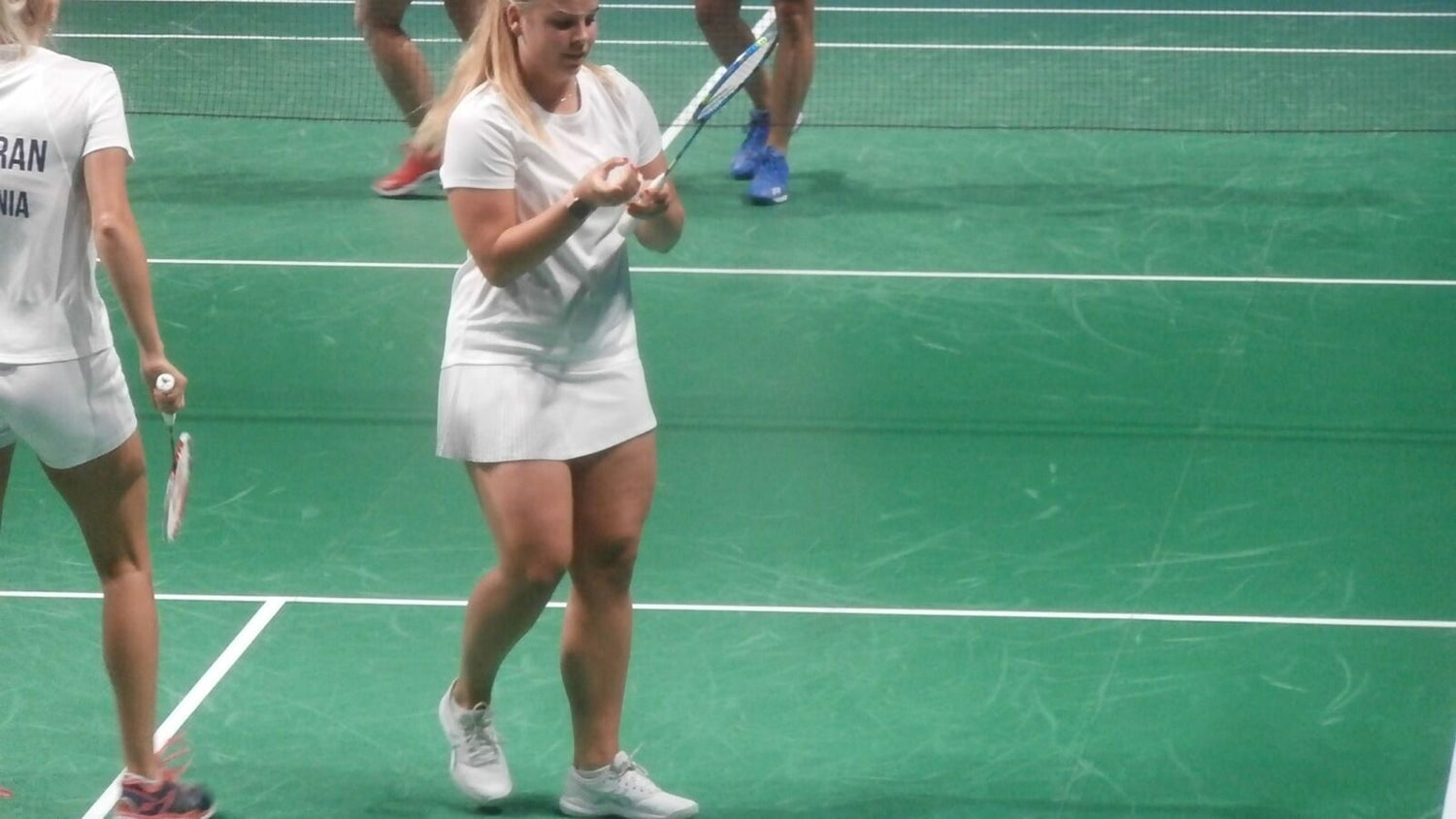 Une personne vêtue de blanc sur un terrain de badminton intérieur tient le manche de la raquette à deux mains près du filet; une partenaire ou un partenaire de double et les chaussures bleues d’un adversaire sont visibles sur le terrain vert marqué.