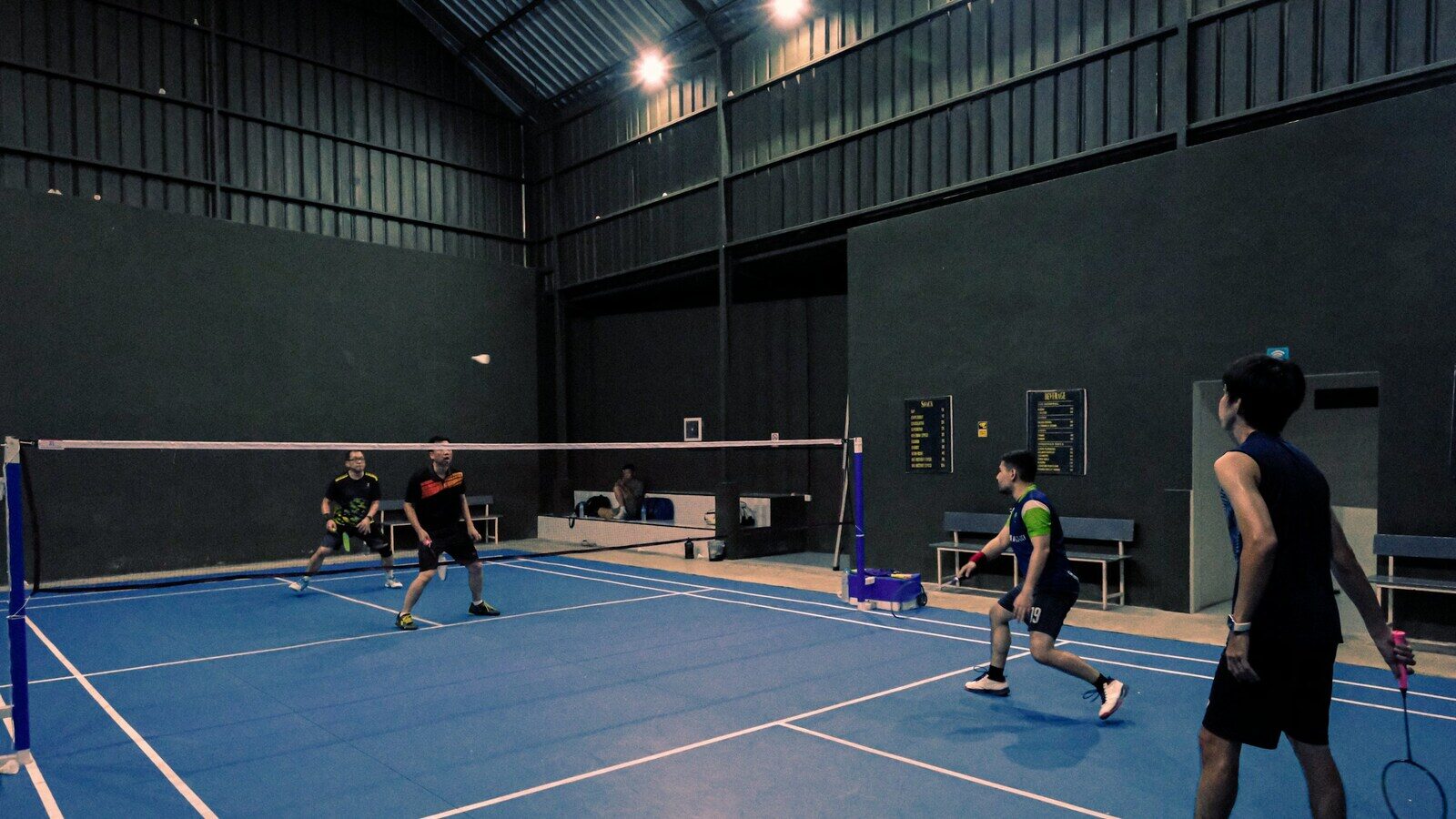 Four club badminton players on an indoor court during a doubles rally, framed above the nearest player's shoes, with the front-court player lunging low to reach a shuttle near the net.