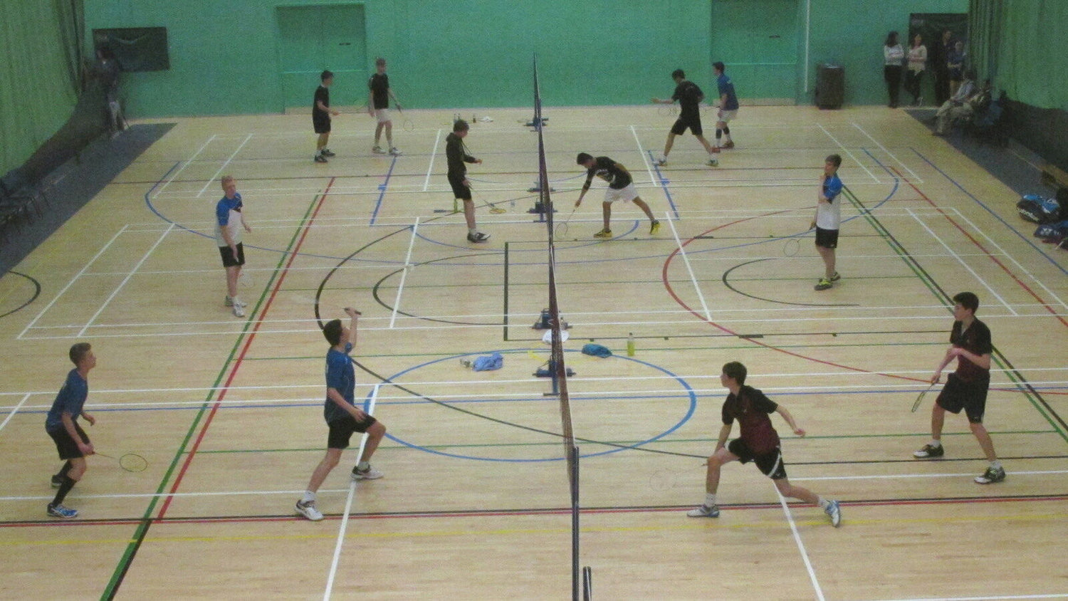 Douze joueurs en paires de double jouent des échanges en même temps sur trois terrains de badminton côte à côte dans un gymnase scolaire, tandis que quelques spectateurs regardent depuis le mur du fond.