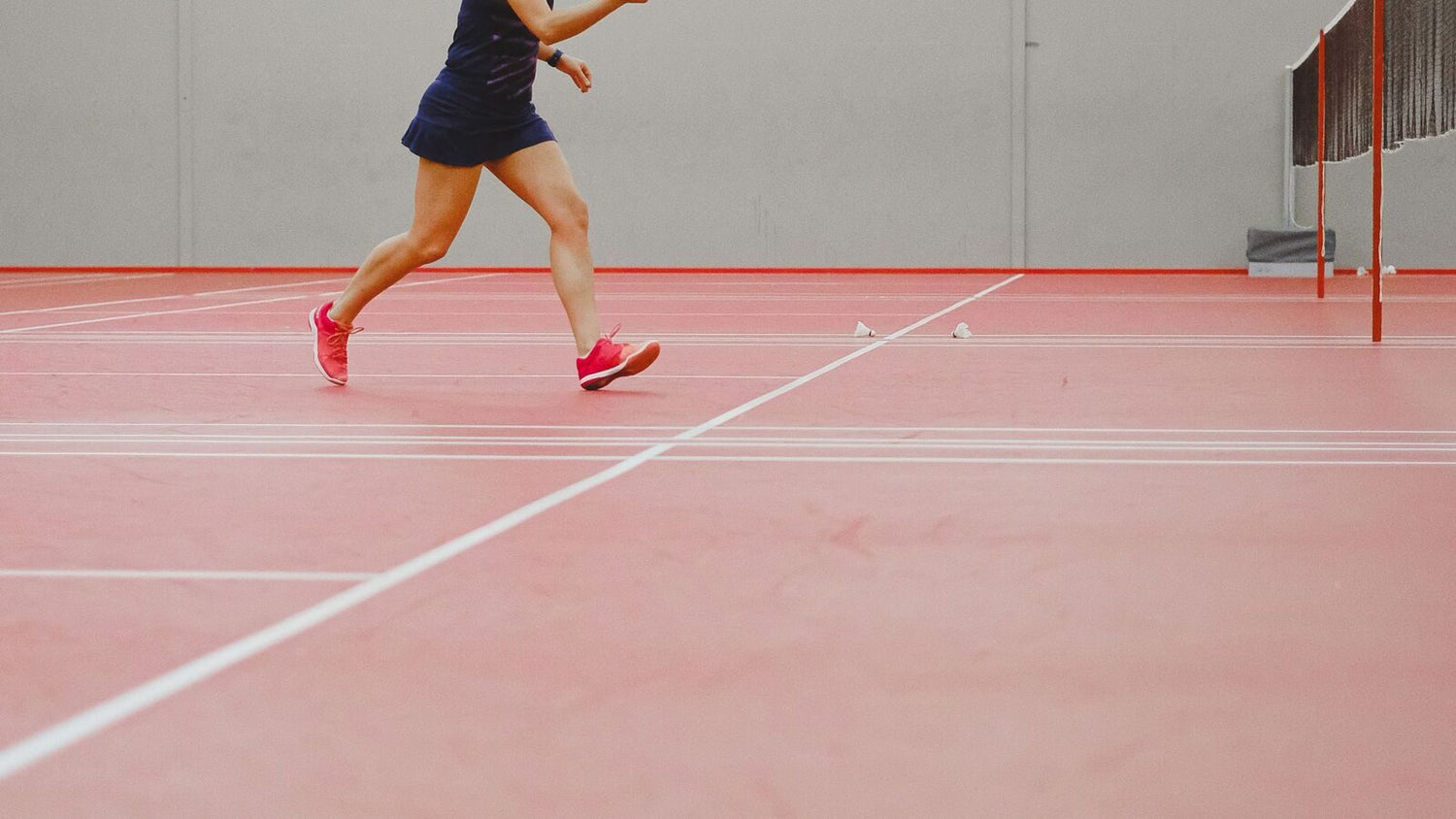 Les jambes et les pieds d’une personne qui joue au badminton au milieu d’une fente sur un terrain intérieur, le pied avant chaussé de rose se posant vers l’extérieur sur la surface rouge, avec le filet et le poteau visibles derrière.