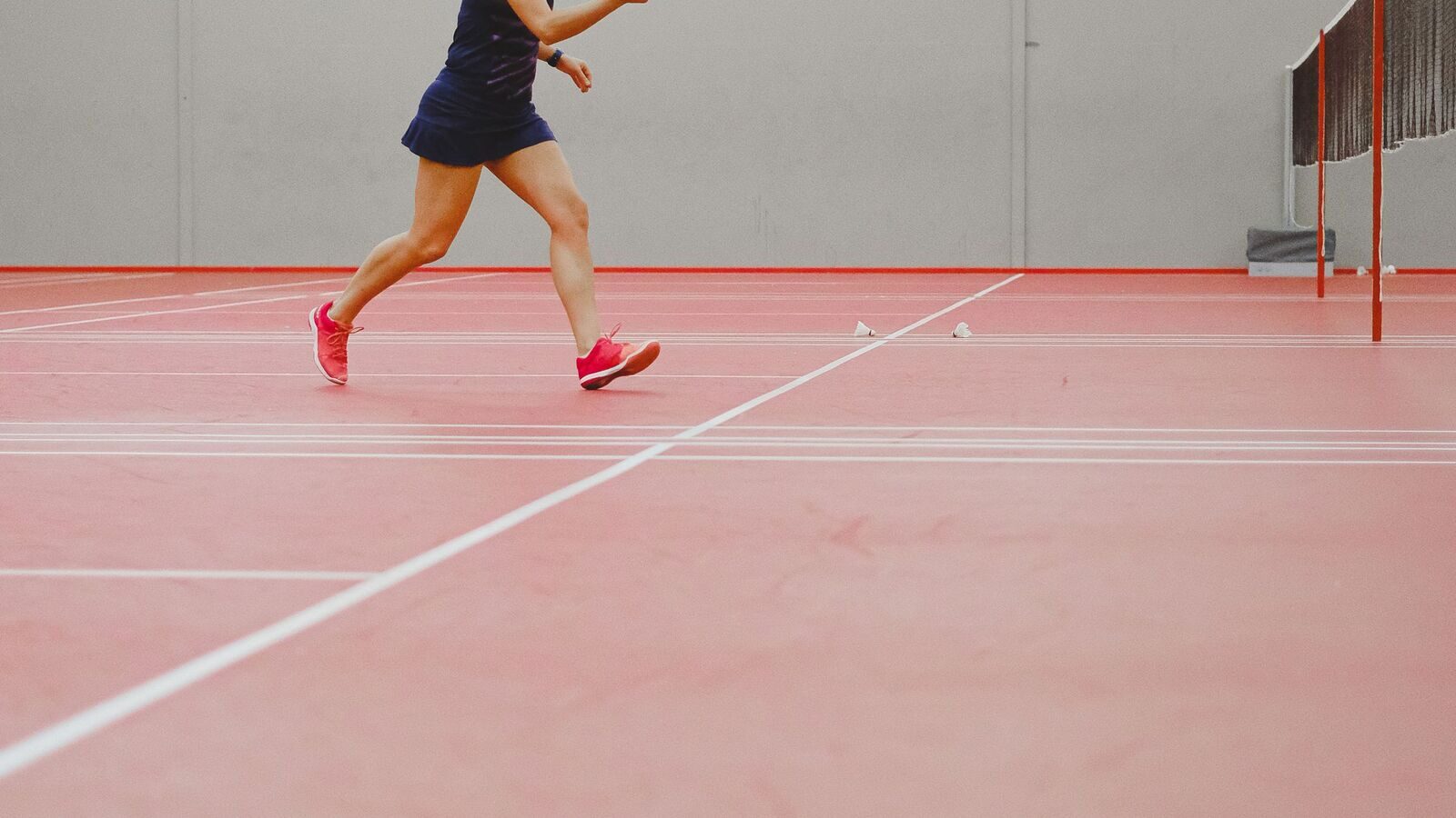 Les jambes et les pieds d’une personne qui joue au badminton au milieu d’une fente sur un terrain intérieur, le pied avant chaussé de rose se posant vers l’extérieur sur la surface rouge, avec le filet et le poteau visibles derrière.