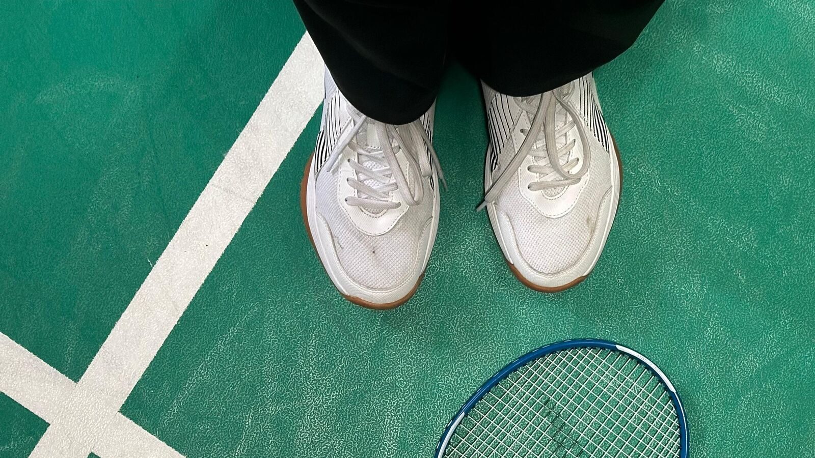 Vue du dessus des chaussures d’une personne qui joue au badminton, debout à côté de sa raquette posée sur la ligne du terrain, dans un moment d’arrêt avant le jeu.