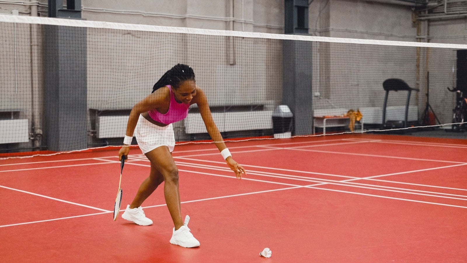 Une personne qui joue au badminton s’élance bas vers un volant sur un terrain intérieur, avec de simples chaussures blanches et la raquette près du sol.