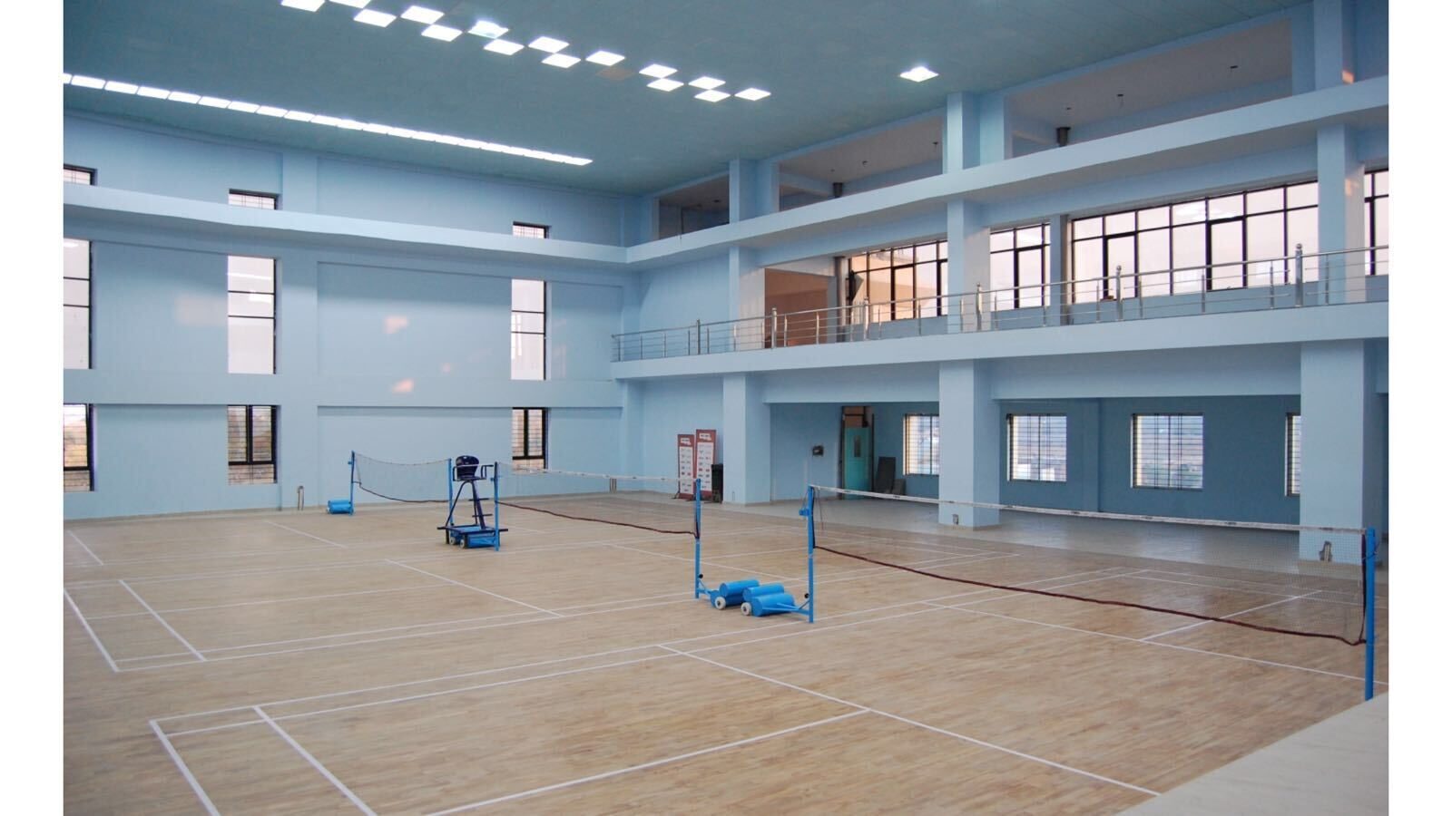 Gymnase intérieur de badminton vide avec deux terrains installés côte à côte, filets en place, chaise d’arbitre sur un terrain et chariot de volants à côté de l’autre, sous une galerie à deux étages.