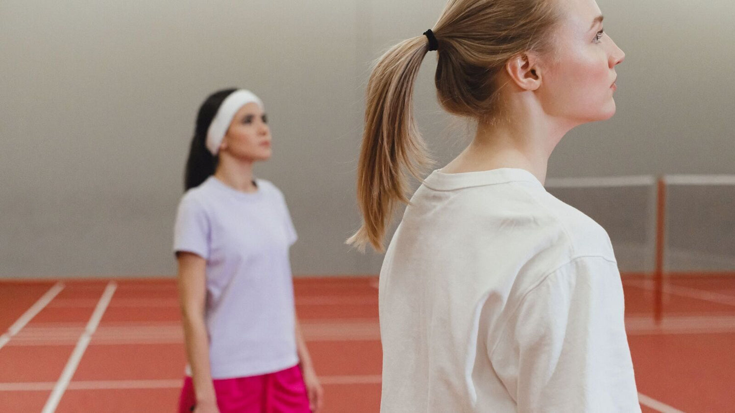 Deux jeunes femmes en vêtements de sport se tiennent de profil sur un terrain de badminton intérieur rouge, tournées vers un filet flou à l’arrière-plan, sans raquette dans le cadre.