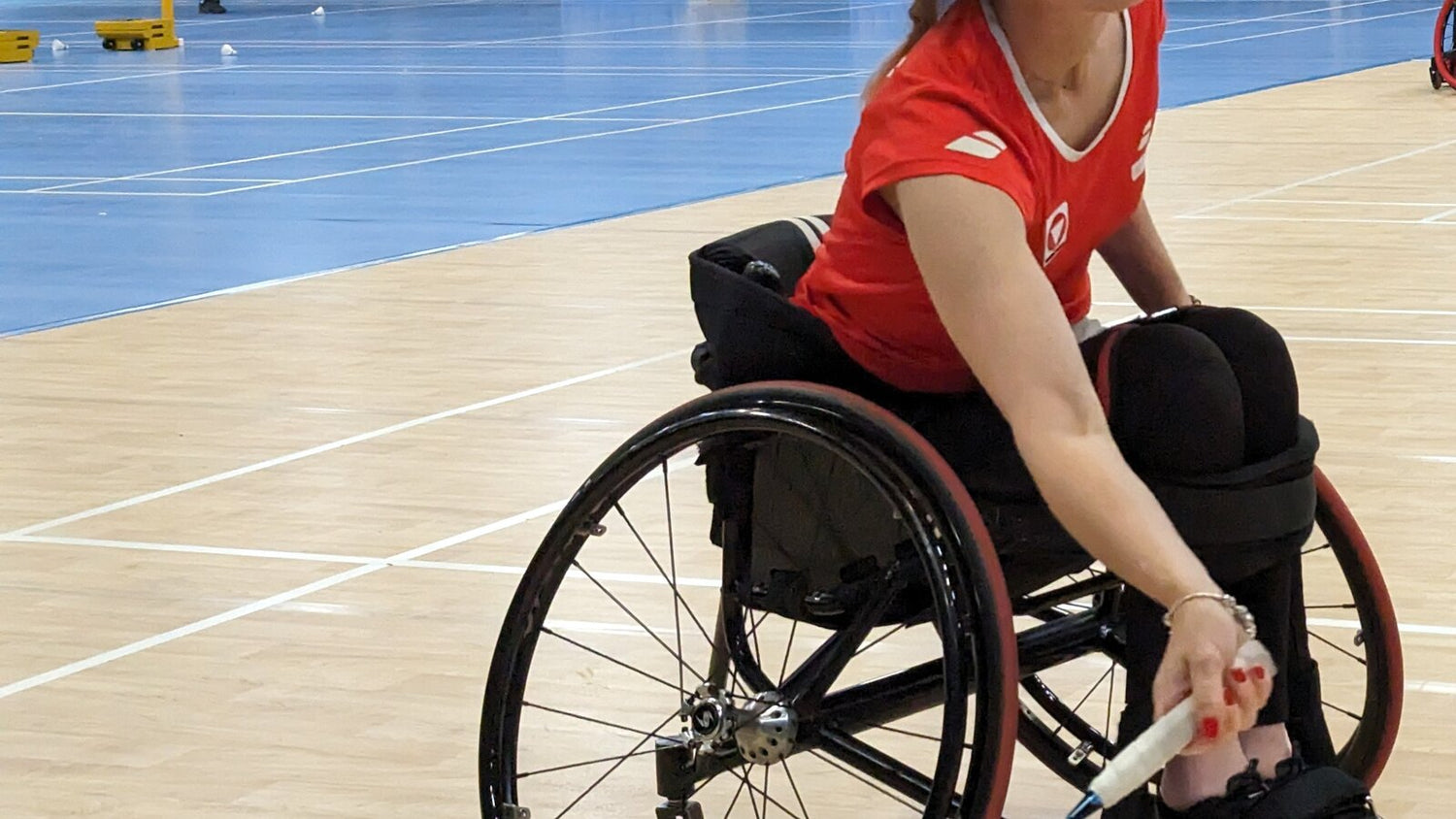Une joueuse de badminton en fauteuil roulant s’étire bas avec sa raquette pendant un entraînement sur un terrain intérieur en bois sur ressorts; les poteaux du filet du terrain adjacent et des volants épars se trouvent derrière elle.