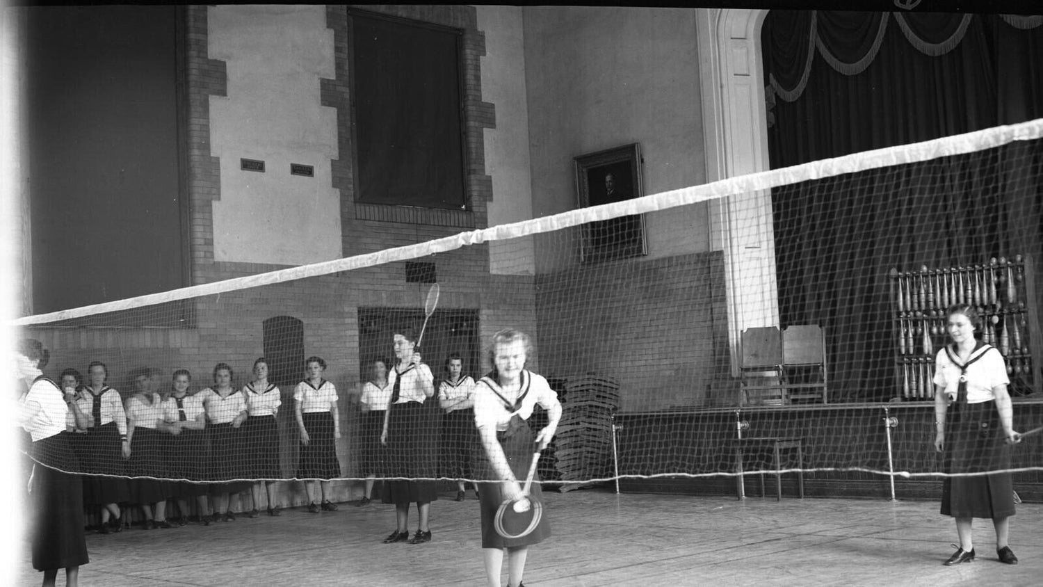 Photo d’archives en noir et blanc datant de 1937 montrant des élèves en uniforme de sport qui jouent au badminton dans un gymnase; deux personnes jouent un échange au filet et une file de coéquipiers attend derrière elles
