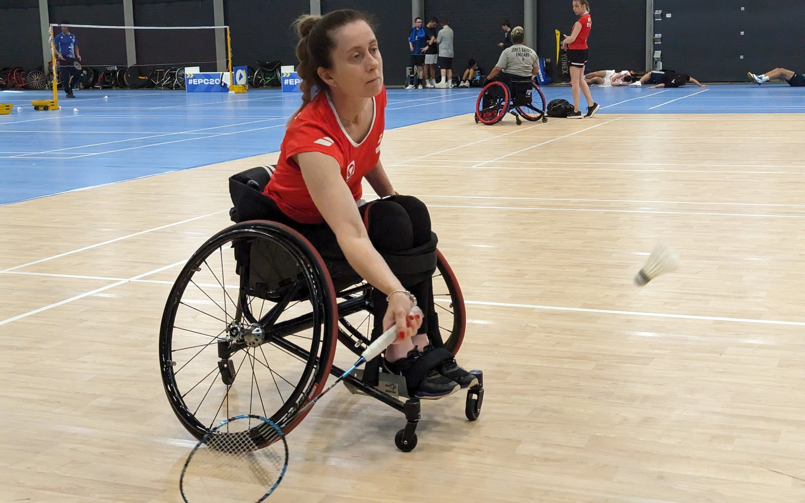 Joueuse de badminton en fauteuil roulant dans un fauteuil de sport, réalisant un revers bas dans une salle d’entraînement avec des terrains et des fauteuils de sport de rechange derrière elle.