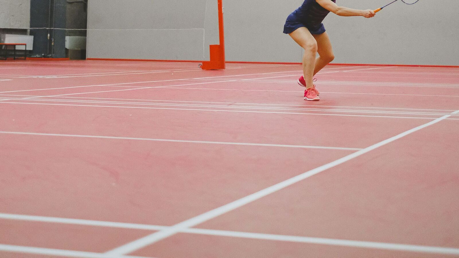 Une personne qui joue au badminton fait une fente vers le filet pour jouer un coup sur un terrain intérieur marqué, avec le poteau du filet et les lignes de service et de côté qui se croisent visibles sur la surface rouge