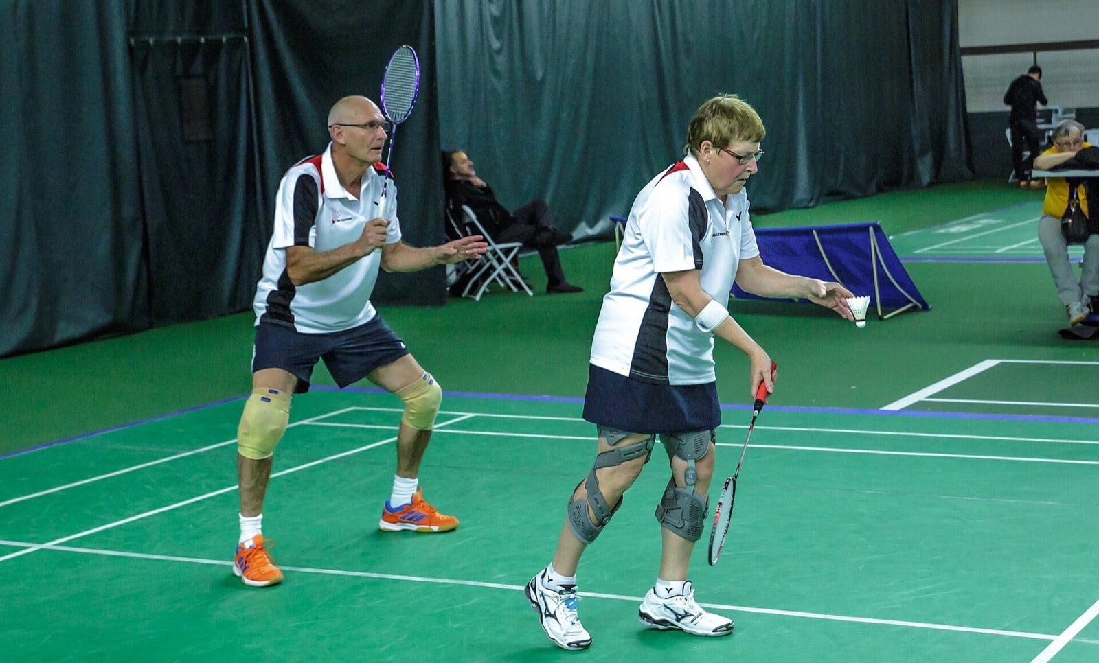 Deux personnes jouant en double chez les maîtres au milieu d’un échange sur un terrain de badminton intérieur, la personne au service portant des genouillères.
