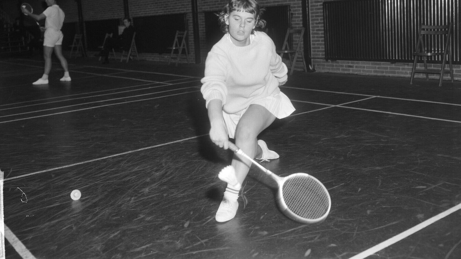 Photo d’archives en noir et blanc de 1965 d’une joueuse de badminton accroupie, abaissant sa raquette en bois en travers du corps pour retourner un volant près du sol; un second volant est près d’elle et une partenaire échange derrière elle.