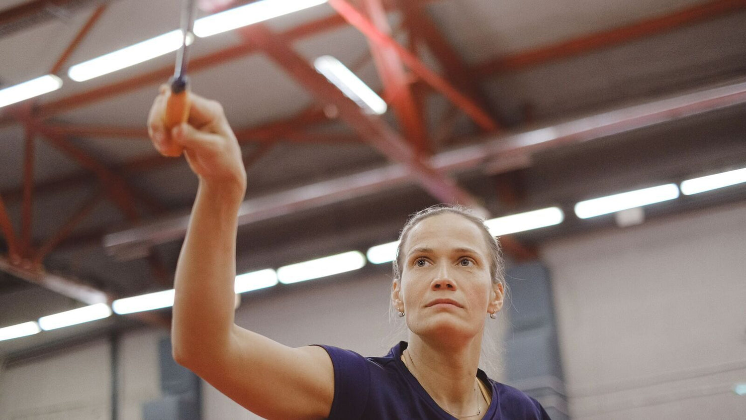 Joueuse sur un terrain de badminton intérieur levant sa raquette droit au-dessus de sa tête, le bras complètement tendu et les yeux suivant le volant vers le haut, en préparation de la frappe.