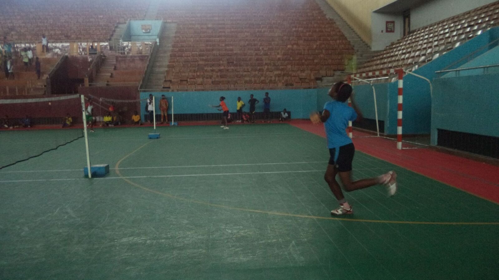 Une jeune joueuse s’élance pour effectuer un smash au-dessus de la tête sur un terrain de badminton intérieur, avec les poteaux du filet et des gradins vides visibles derrière elle.