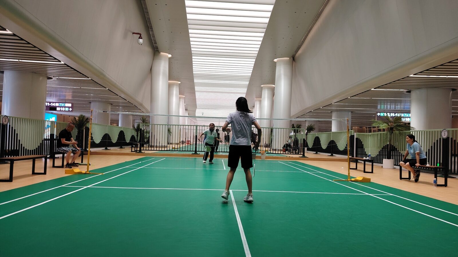 Des joueurs disputent un échange sur un terrain de badminton intérieur dans une salle sportive de gare, avec un deuxième terrain, des bancs et des spectateurs visibles de chaque côté du filet.