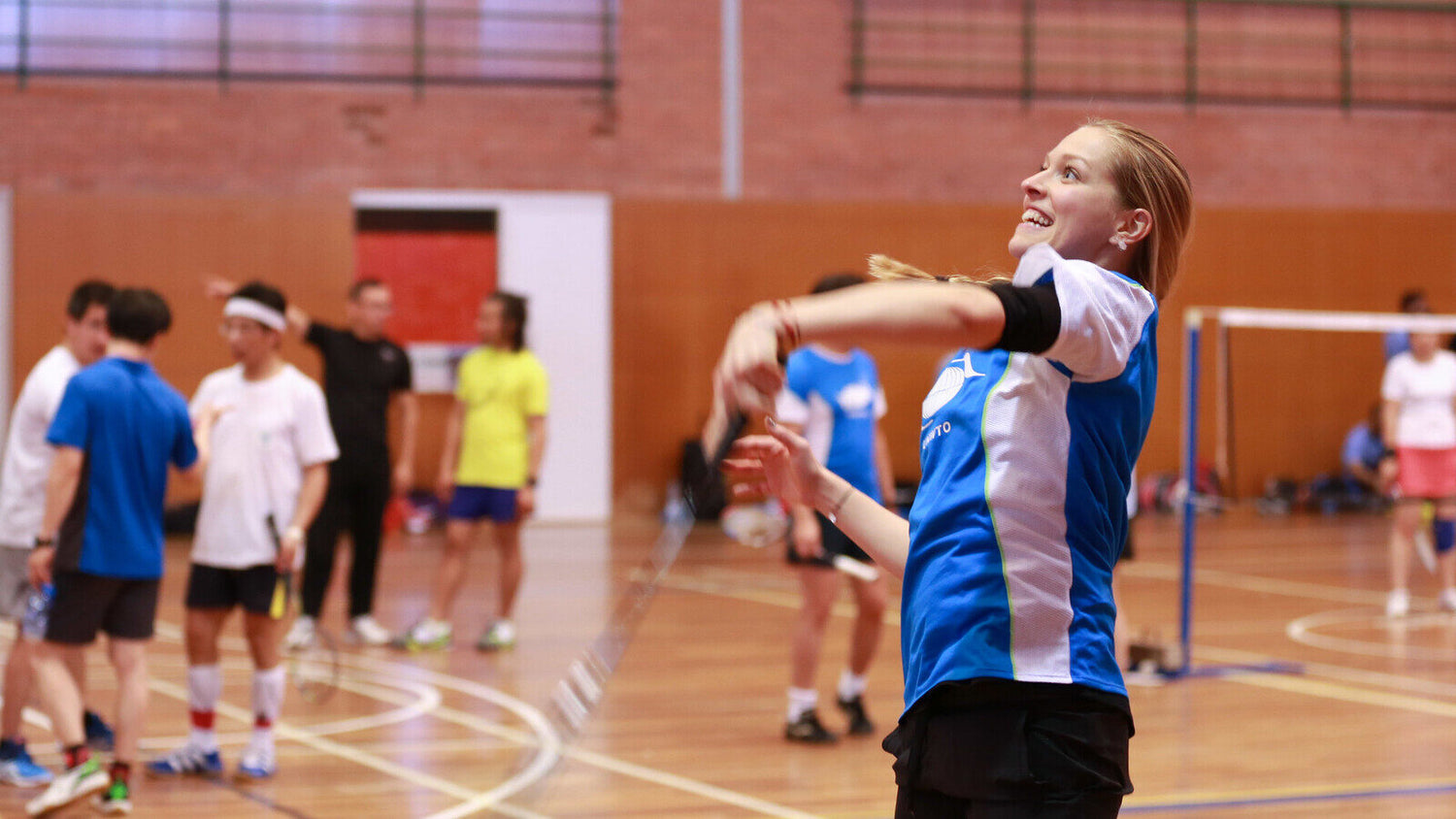 Personne souriante en tenue d’équipe de badminton qui accompagne un coup au-dessus de la tête sur un terrain intérieur, avec une partenaire ou un partenaire de double, des adversaires et le filet visibles à proximité.