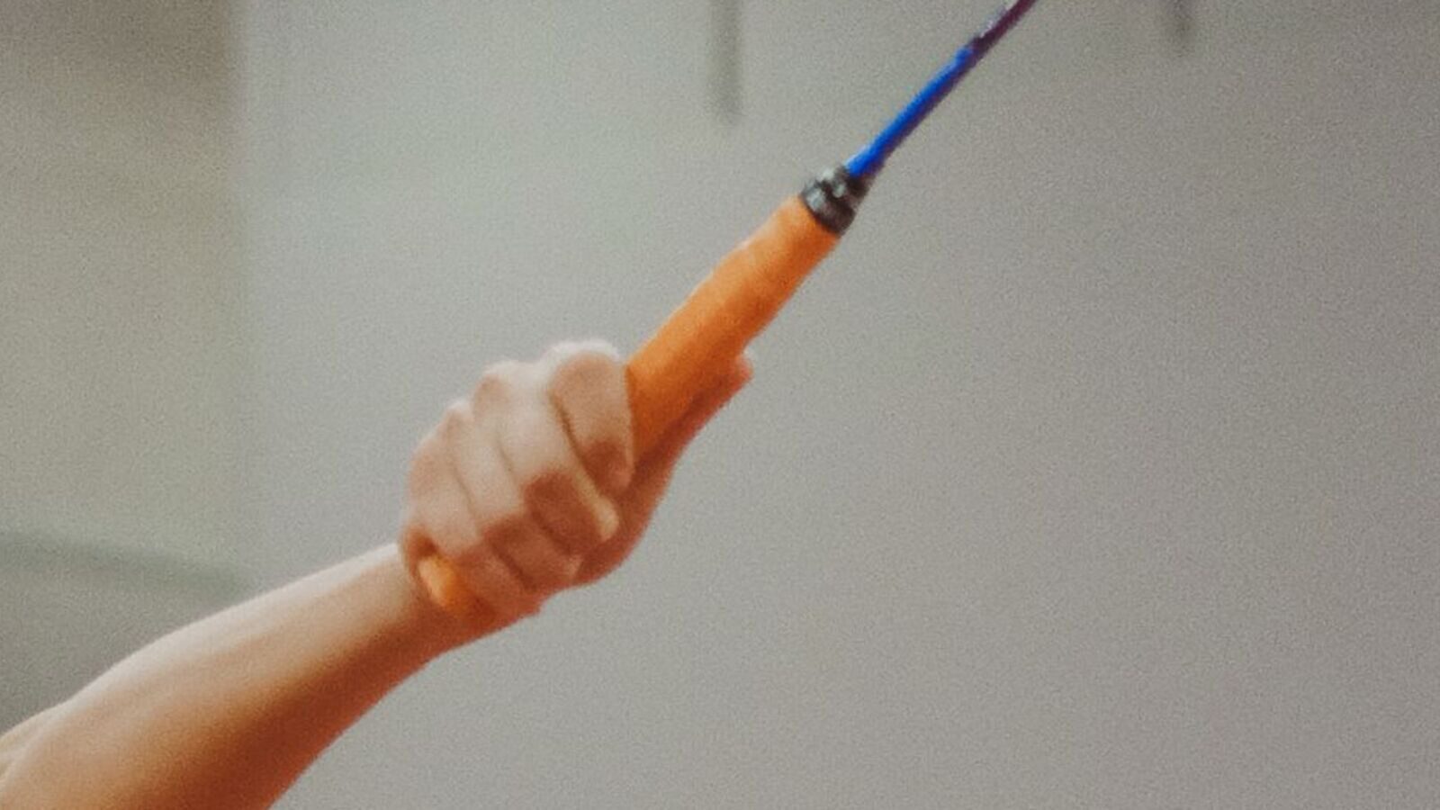 Vue rapprochée de la main d’une personne qui tient une raquette de badminton, le bras tendu vers le haut et l’avant au milieu d’un geste, devant un mur de gymnase uni.