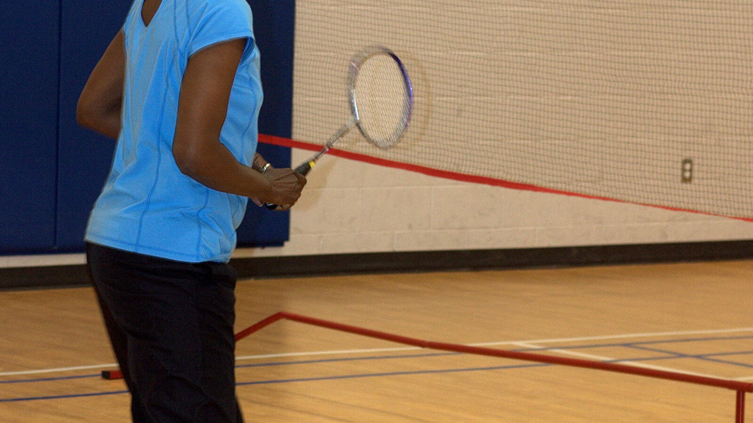 Gros plan sur les bras d’une personne qui tient une raquette de badminton en position d’attente près d’un filet intérieur, avec le terrain et la protection murale visibles derrière.
