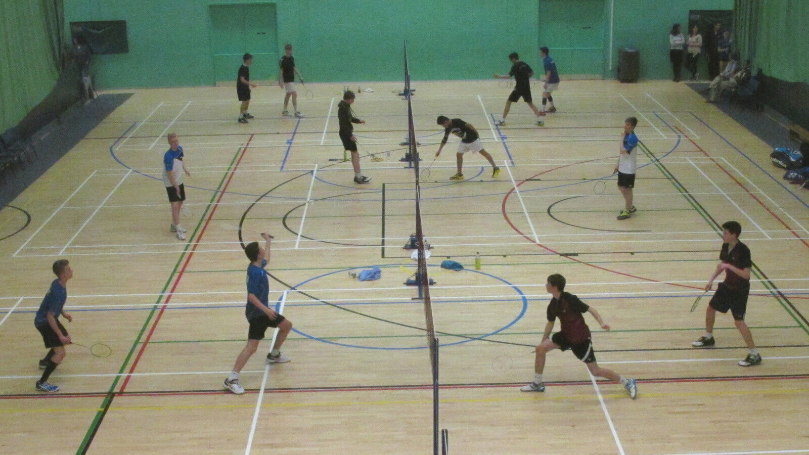 Douze joueurs en paires de double jouent des échanges en même temps sur trois terrains de badminton côte à côte dans un gymnase scolaire, tandis que quelques spectateurs regardent depuis le mur du fond.