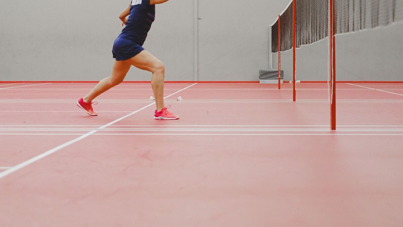 Jambes et torse d’un joueur de badminton en fente sur un terrain intérieur : genou avant très fléchi, jambe arrière tendue, chaussures rouges près de deux volants sur terrain rouge à lignes de service blanches, filet et poteau derrière.