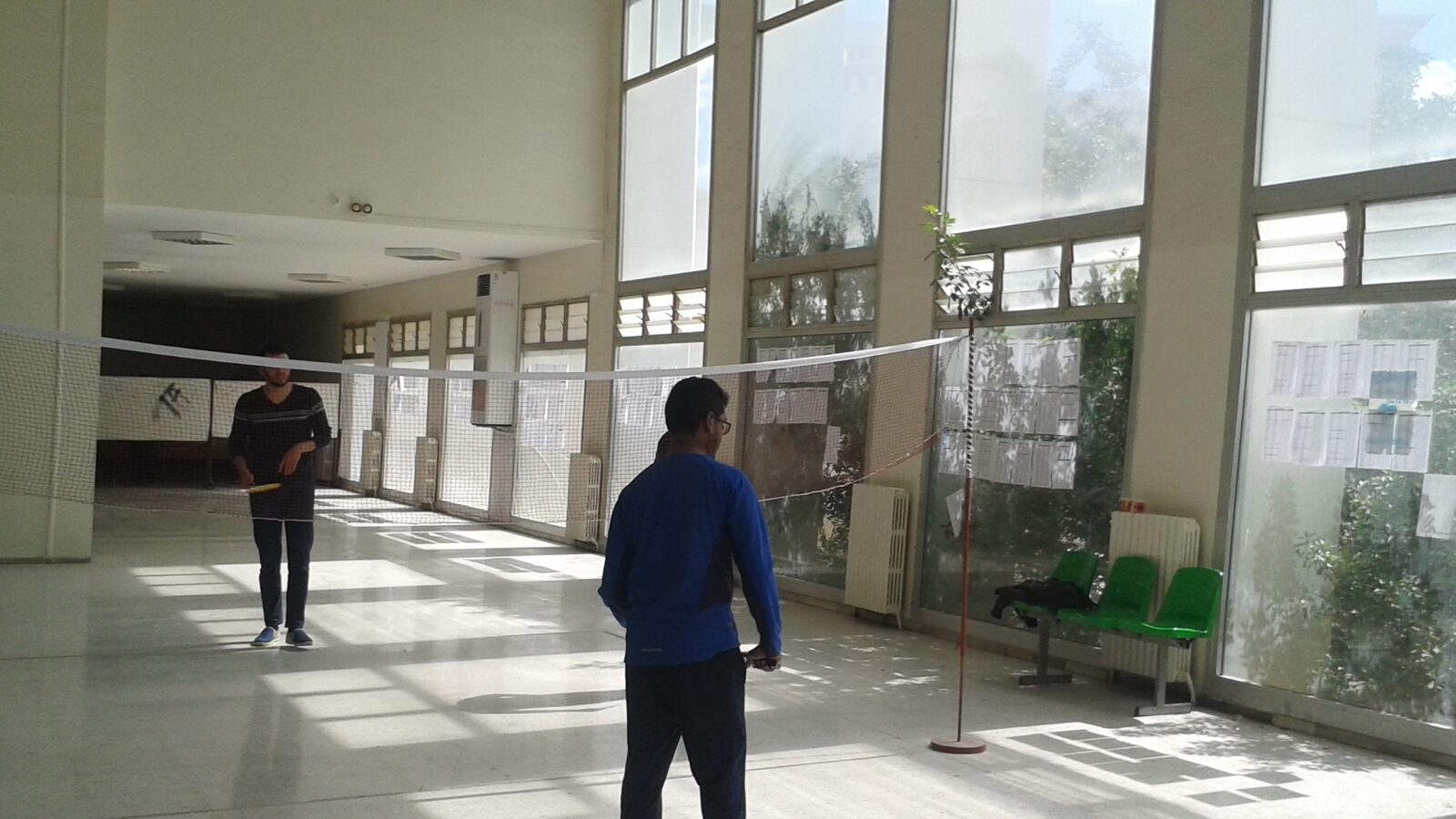 Deux personnes se font face de part et d’autre d’un filet de badminton installé dans un corridor intérieur, l’une tenant une raquette prête à retourner.