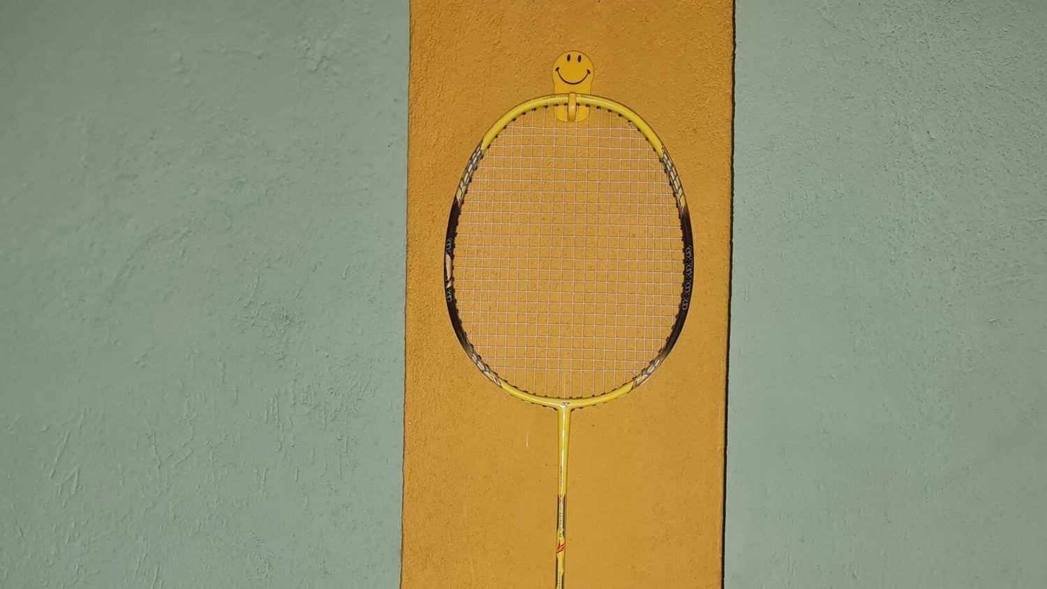 A yellow badminton racket hangs on a hook, showing its full oval head and crossed string bed against a colourful wall.