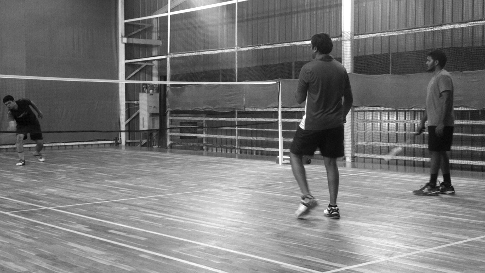 Photo en noir et blanc d’un échange de double de badminton en salle : une personne est accroupie près du filet au fond du terrain tandis que deux personnes se tiennent prêtes près du filet au premier plan, avec un volant visible en plein vol.