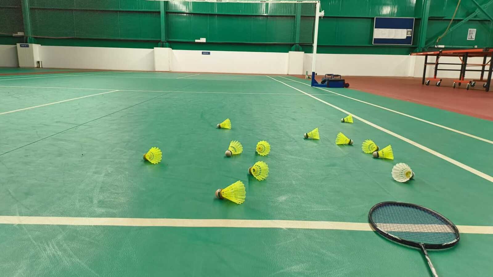 Terrain intérieur de badminton vide entre deux manches, avec une douzaine de volants jaunes en plumes et une raquette éparpillés sur le terrain vert devant le filet et son poteau; lignes de côté du double et chariot d’équipement pliant visibles.