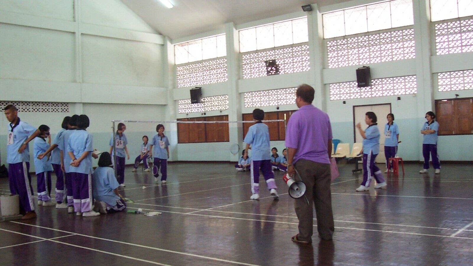 Une personne entraîneuse dirige un groupe d’élèves en survêtements assortis pendant un entraînement d’équipe de badminton en salle, raquettes en main près du filet.