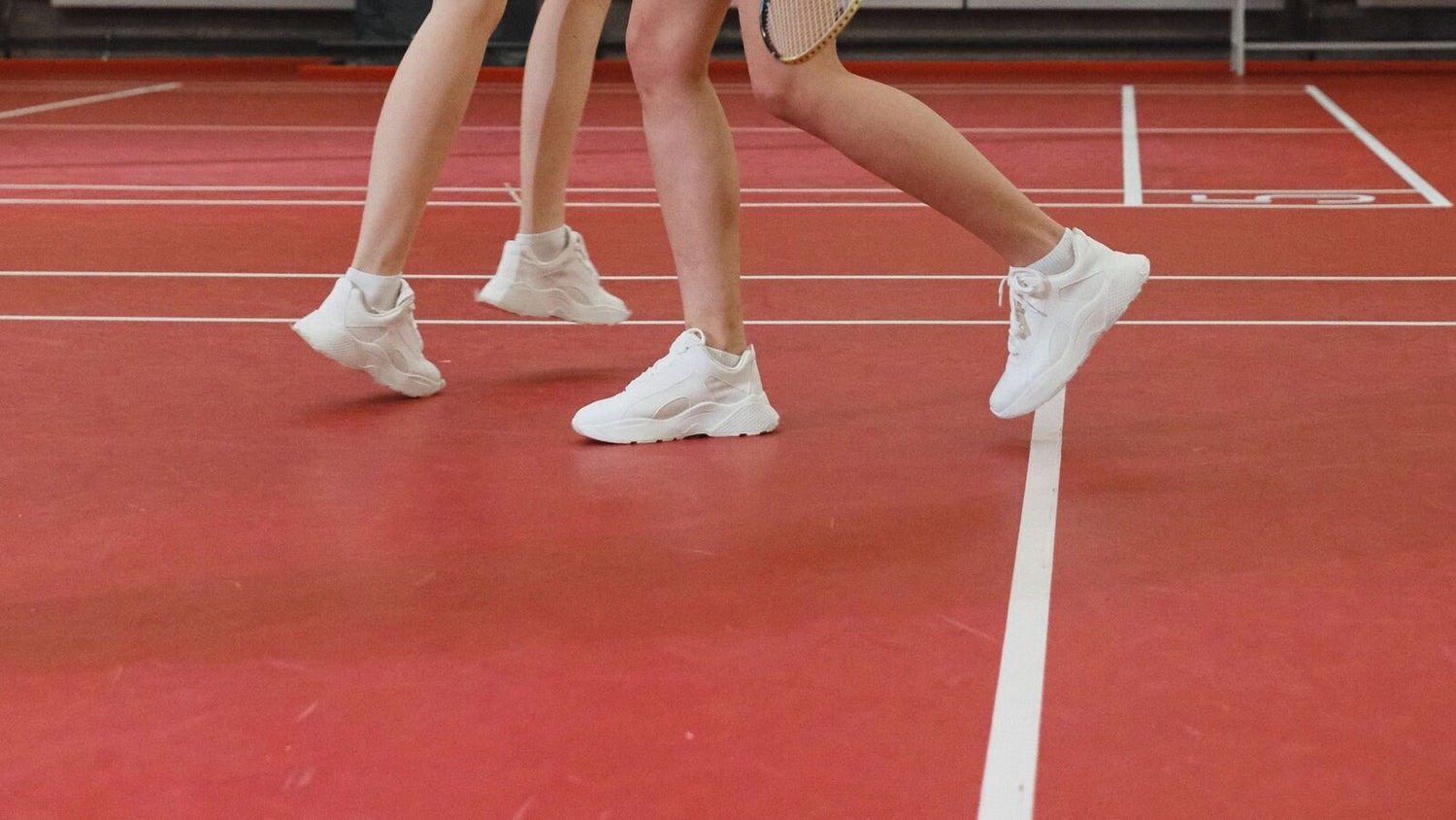 Gros plan sur les jambes de trois personnes jouant au badminton et leurs chaussures de sport blanches, en pleine foulée sur un terrain intérieur rouge, avec une raquette visible au bord de l’image.