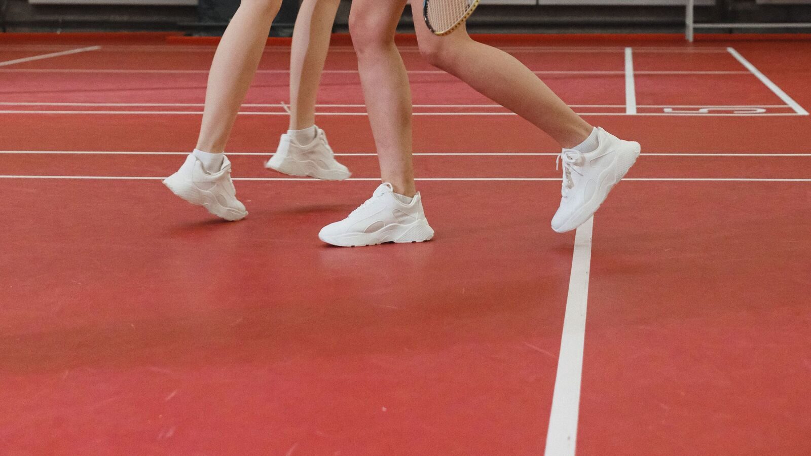 Gros plan sur les jambes de trois personnes jouant au badminton et leurs chaussures de sport blanches, en pleine foulée sur un terrain intérieur rouge, avec une raquette visible au bord de l’image.