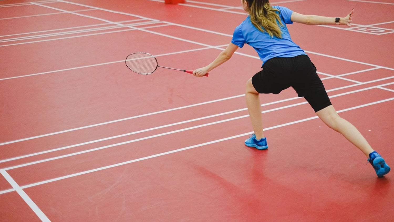 Joueuse de badminton vue de dos qui se fend pour retourner un volant, le bras de raquette tendu à hauteur d’épaule sur un terrain intérieur rouge.