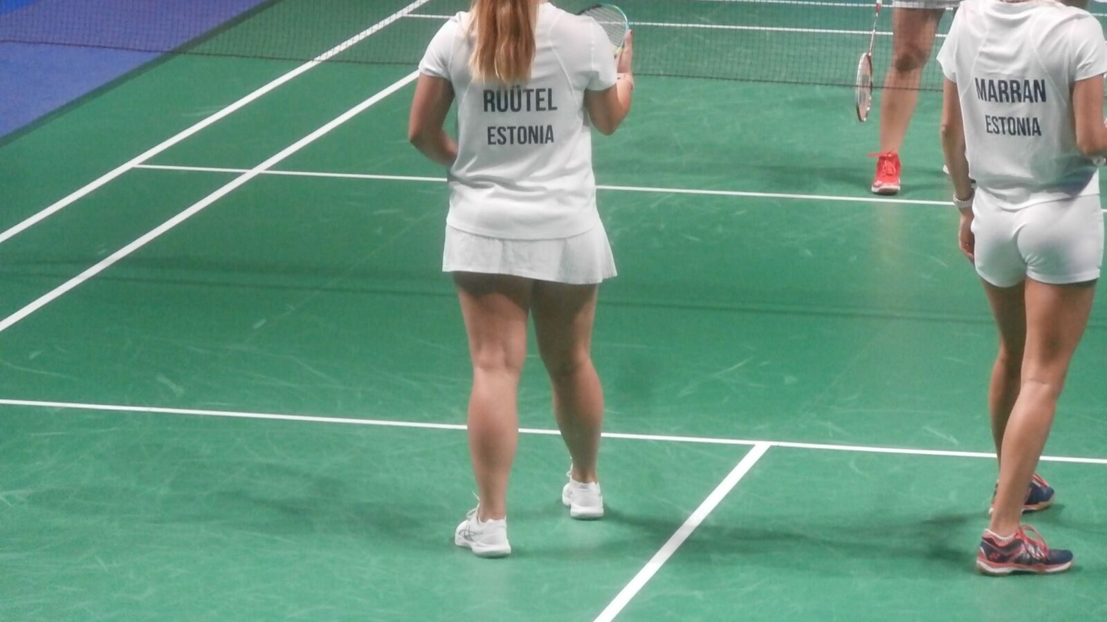Deux joueurs de double se tiennent prêts près du filet sur un terrain de badminton intérieur, dont les lignes de délimitation sont clairement marquées sur la surface verte.