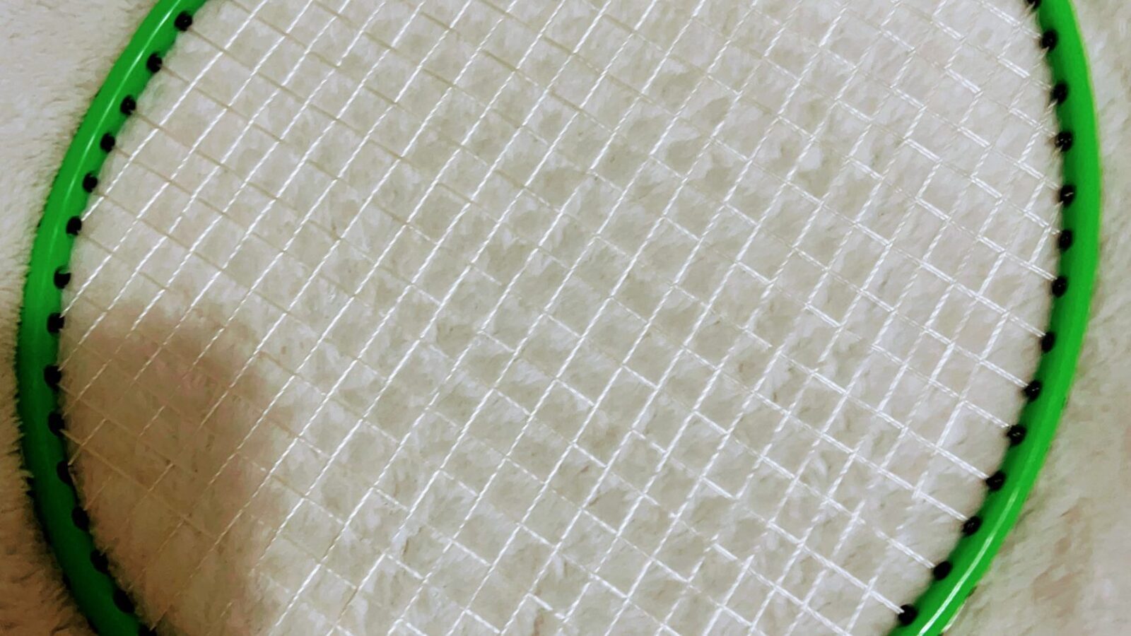 Gros plan sur le cordage d’une raquette de badminton, montrant la grille diagonale des cordes et les trous d’œillets autour du cadre vert.