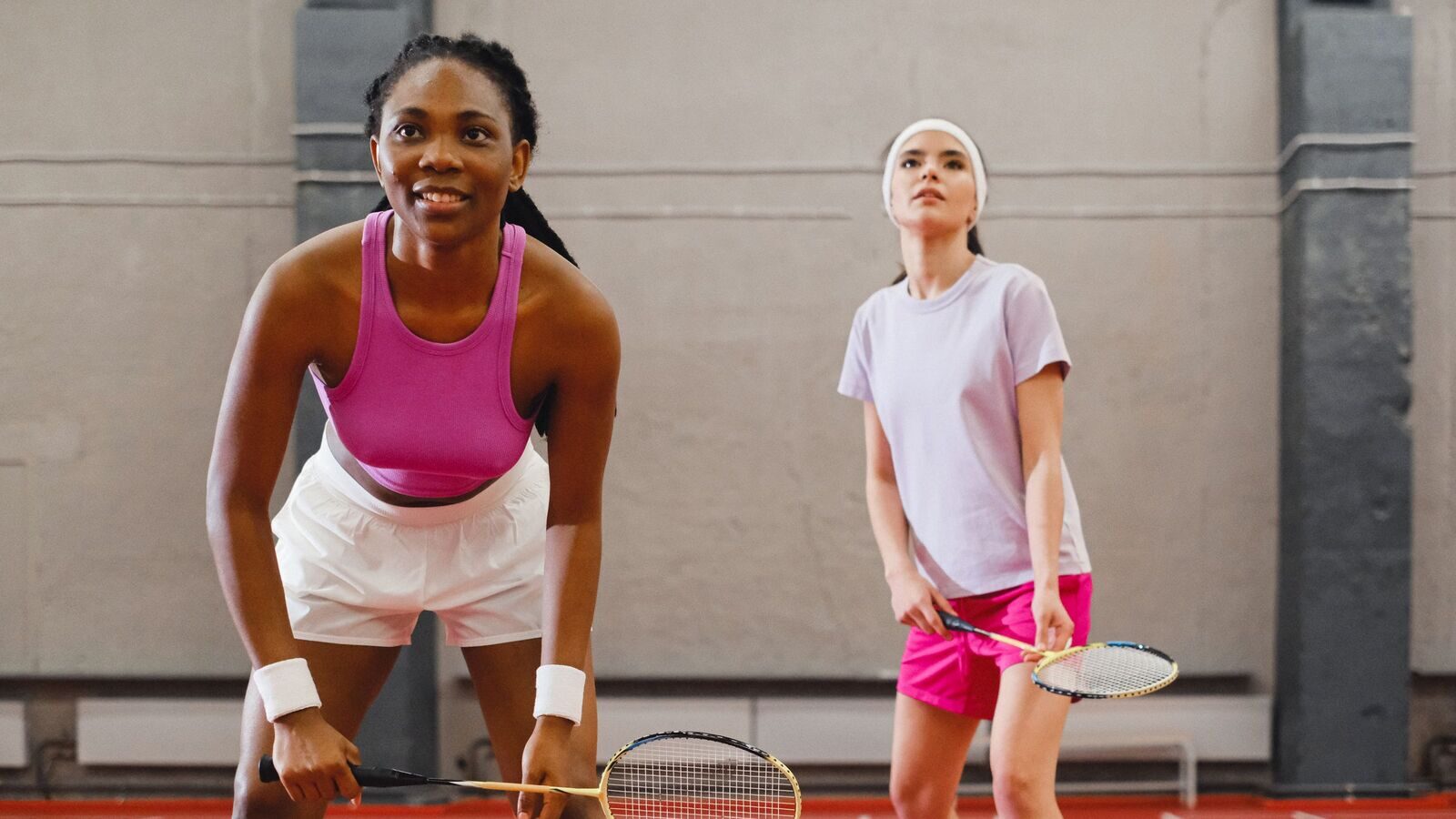 Une joueuse de badminton se repose, les mains sur les genoux, entre deux échanges pendant que sa partenaire de double, portant un bandeau, suit le volant; toutes deux tiennent leur raquette sur un terrain intérieur.