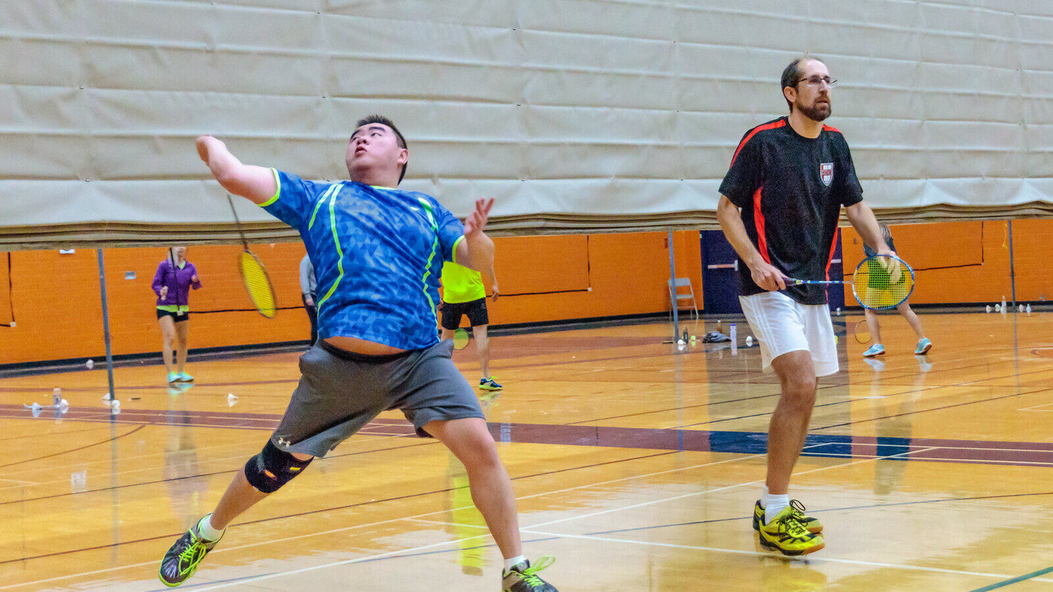 Deux joueurs de double récréatif de badminton sur un terrain intérieur en bois : l’un fait une fente, raquette levée au-dessus de la tête pour un smash; son partenaire est en position d’attente, d’autres joueurs s’entraînent sur les terrains derrière.