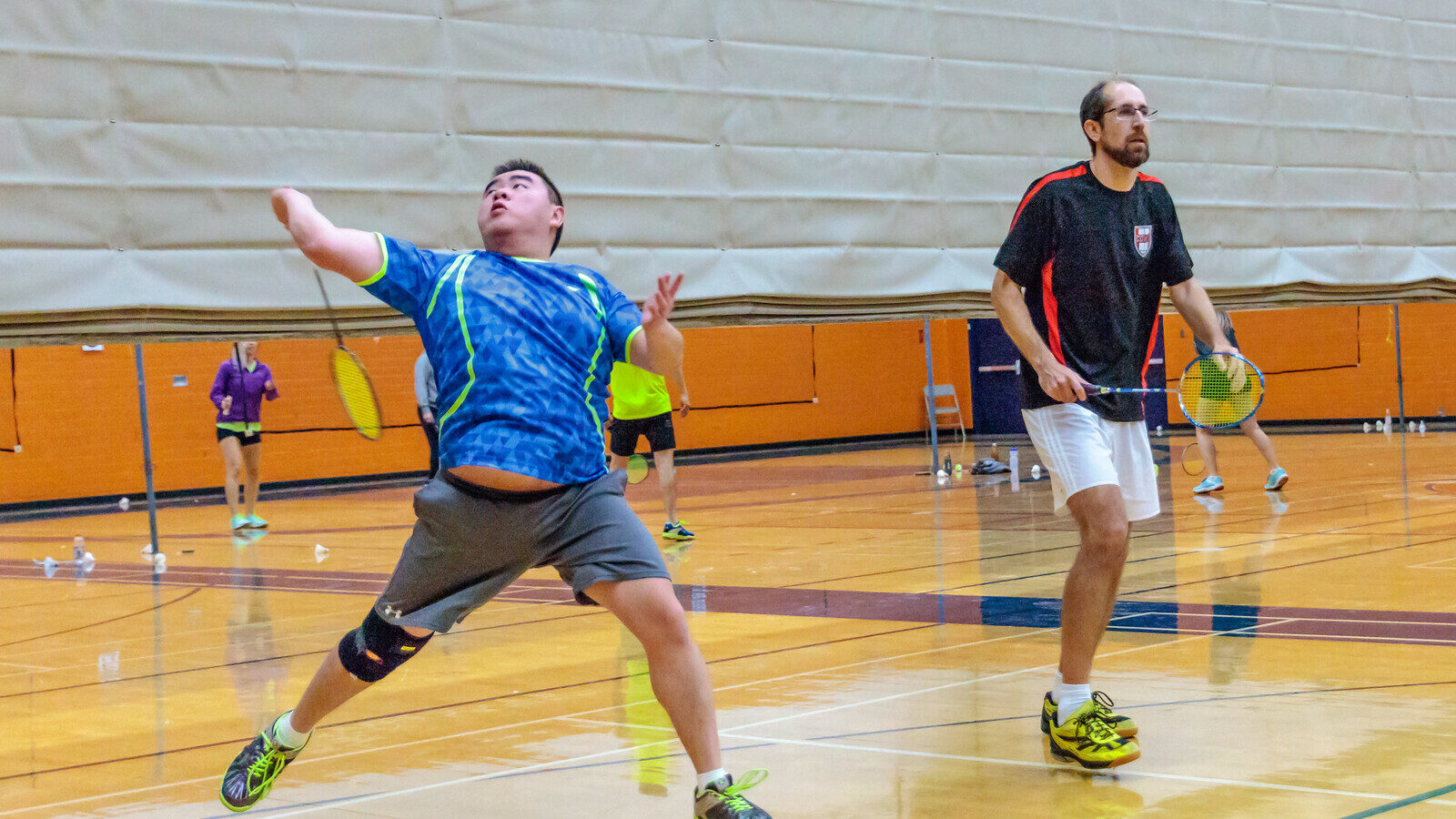 Deux joueurs de double récréatif de badminton sur un terrain intérieur en bois : l’un fait une fente, raquette levée au-dessus de la tête pour un smash; son partenaire est en position d’attente, d’autres joueurs s’entraînent sur les terrains derrière.