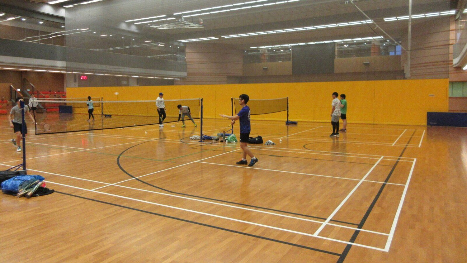 Plusieurs terrains de badminton côte à côte dans une salle de sport intérieure, avec des paires de personnes qui s’échauffent sur chaque terrain avant un match par équipes.