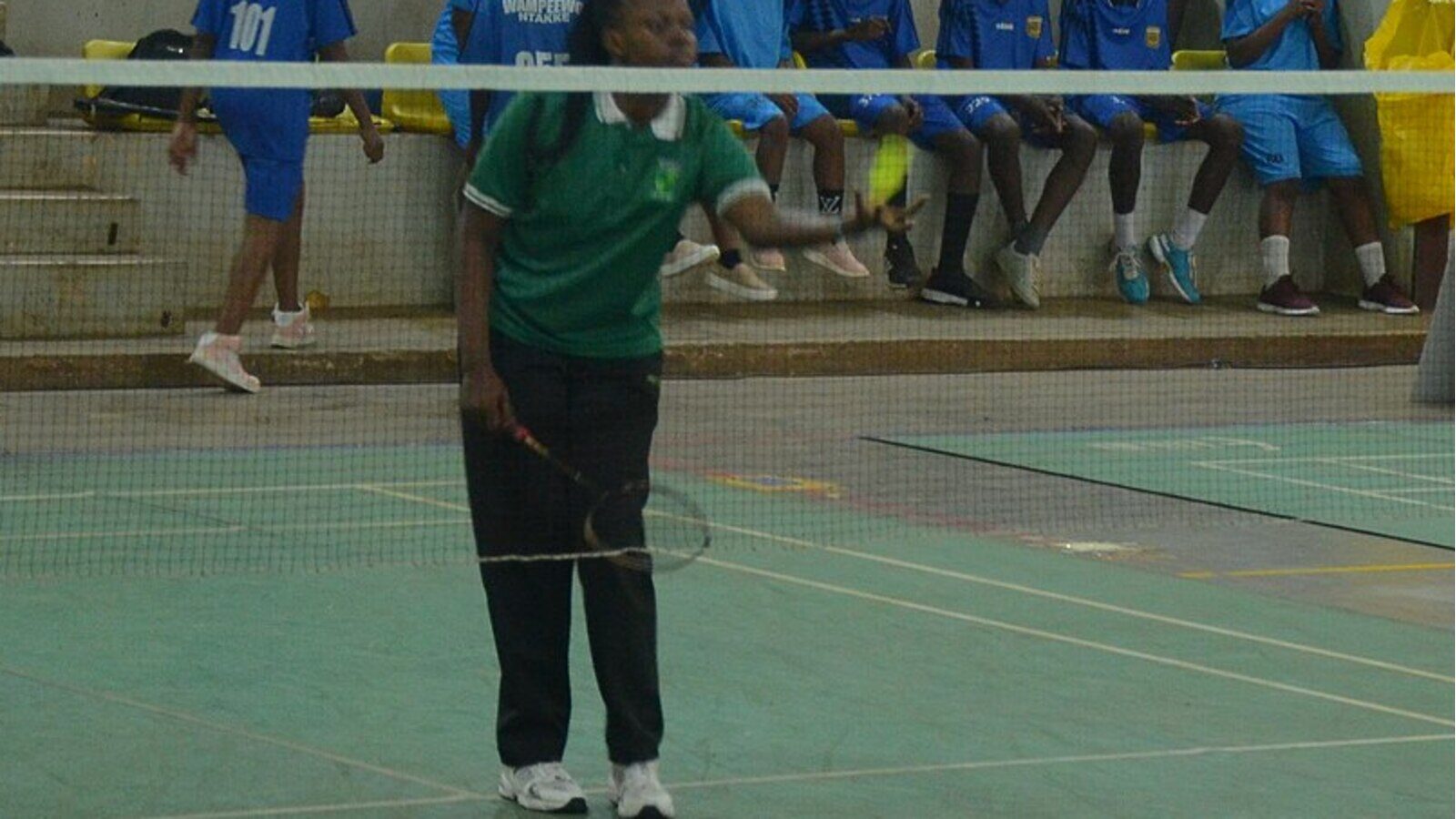 Dans un gymnase scolaire intérieur, une joueuse tient une raquette de badminton et un volant jaune près du filet, sur le point de servir, tandis que ses coéquipières sont assises dans les gradins derrière elle