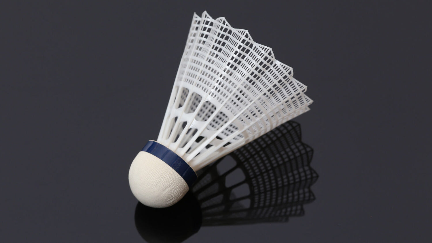 Gros plan sur un volant de badminton blanc à plumes, debout sur une surface sombre réfléchissante; sa jupe nervurée et sa base arrondie sont éclairées sur fond d’ombre.