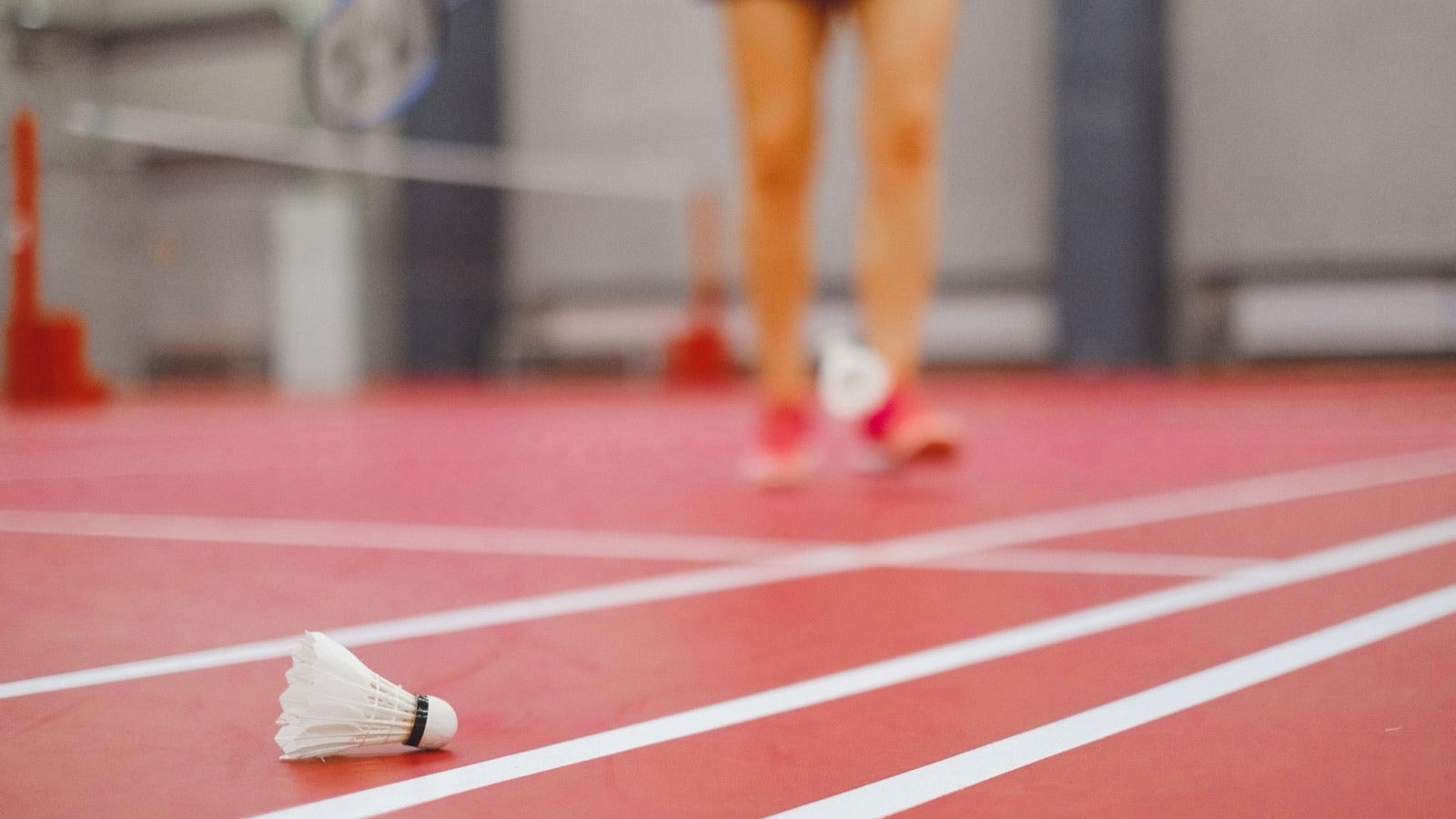 Un volant de badminton repose exactement sur la ligne de délimitation d’un terrain intérieur, tandis que les jambes floues et la raquette d’une personne qui joue s’approchent à l’arrière-plan.