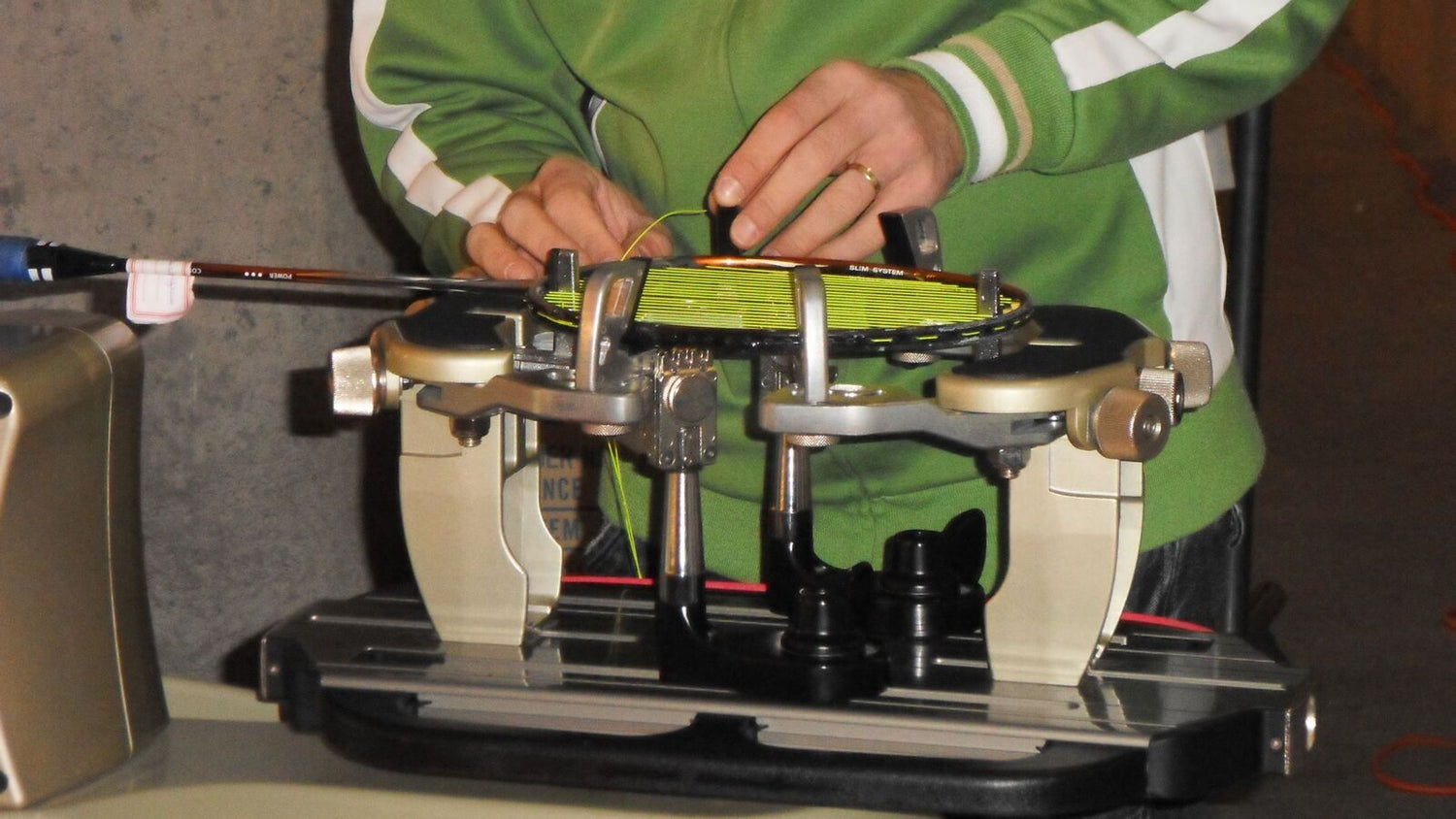 Une paire de mains tisse une corde sur une raquette de badminton fixée dans une machine à cordage à six points, dans un atelier intérieur.