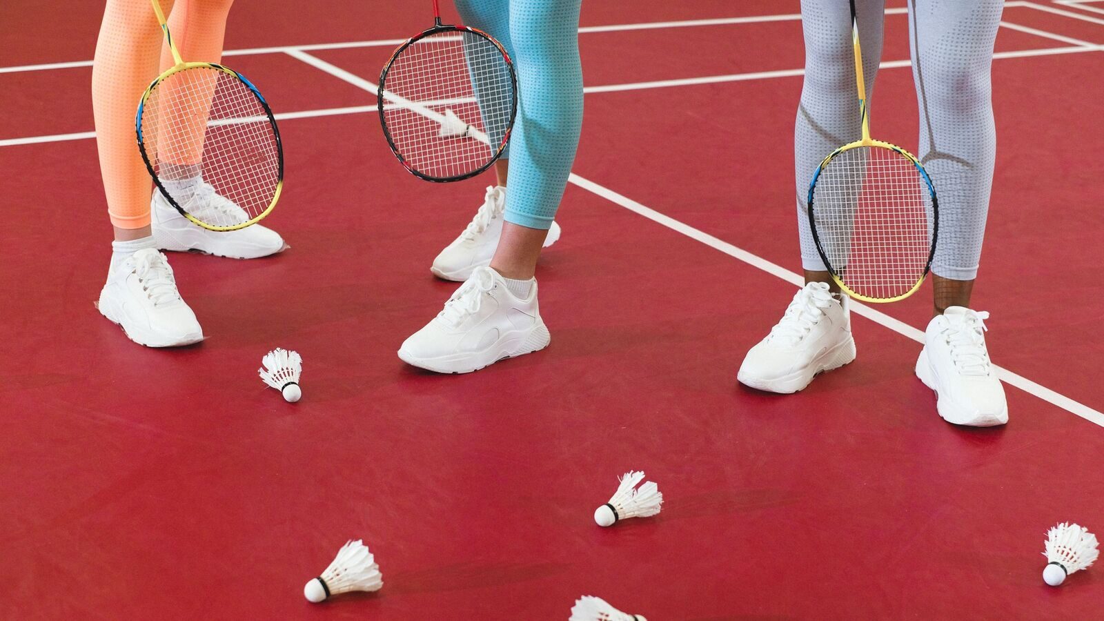 Jambes et têtes de raquettes de trois personnes qui jouent au badminton sur un terrain intérieur rouge, avec plusieurs volants éparpillés sur le sol.