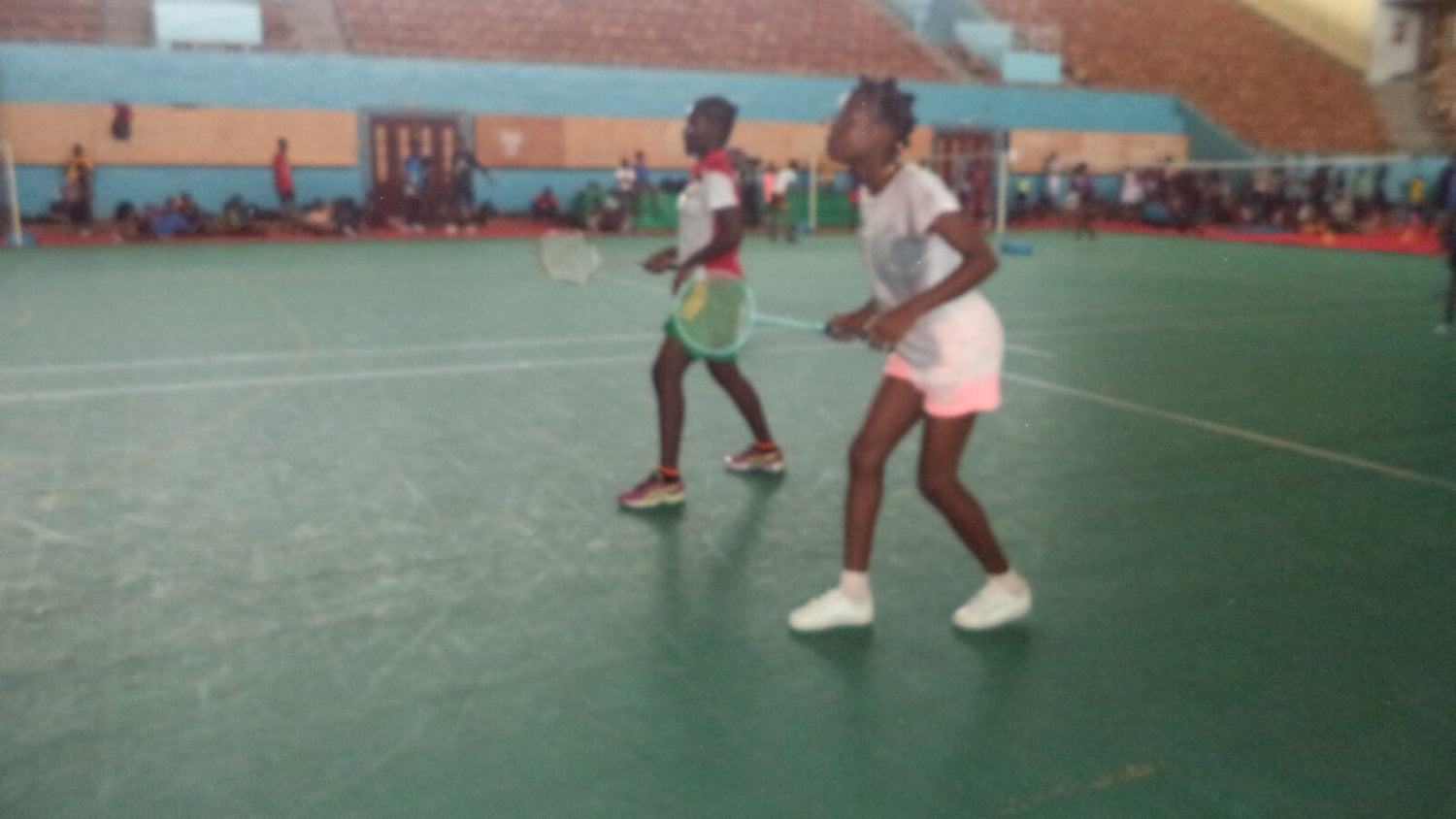 Deux jeunes joueurs en position de double sur un terrain de badminton intérieur, raquettes levées et volant en vol entre eux, avec des gradins vides en arrière-plan.
