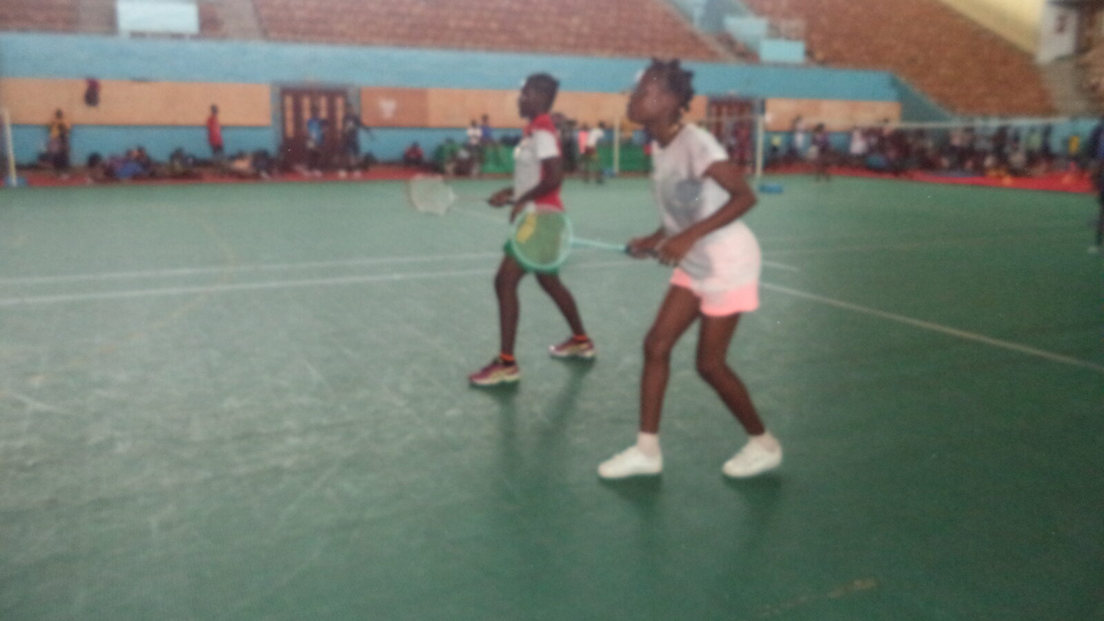 Deux jeunes joueurs en position de double sur un terrain de badminton intérieur, raquettes levées et volant en vol entre eux, avec des gradins vides en arrière-plan.