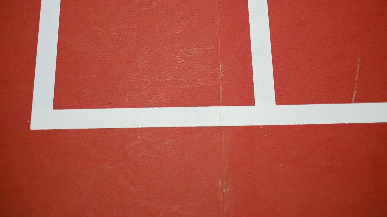 Gros plan sur les lignes blanches d’un terrain de badminton intérieur rouge, à la jonction en L où la ligne de service court rencontre la ligne centrale ou la ligne de côté du couloir de simple sur demi-terrain.