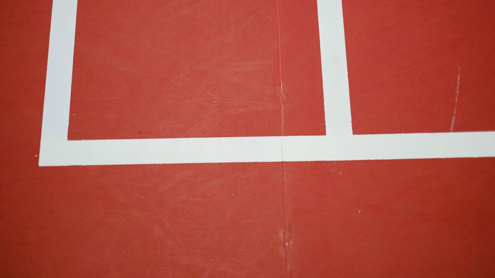 Gros plan sur les lignes blanches d’un terrain de badminton intérieur rouge, à la jonction en L où la ligne de service court rencontre la ligne centrale ou la ligne de côté du couloir de simple sur demi-terrain.