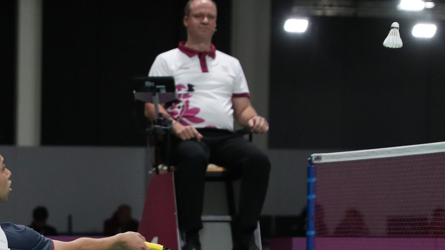 Une personne arbitre de badminton est assise sur la chaise surélevée près du filet et observe un échange pendant que le volant passe devant elle.